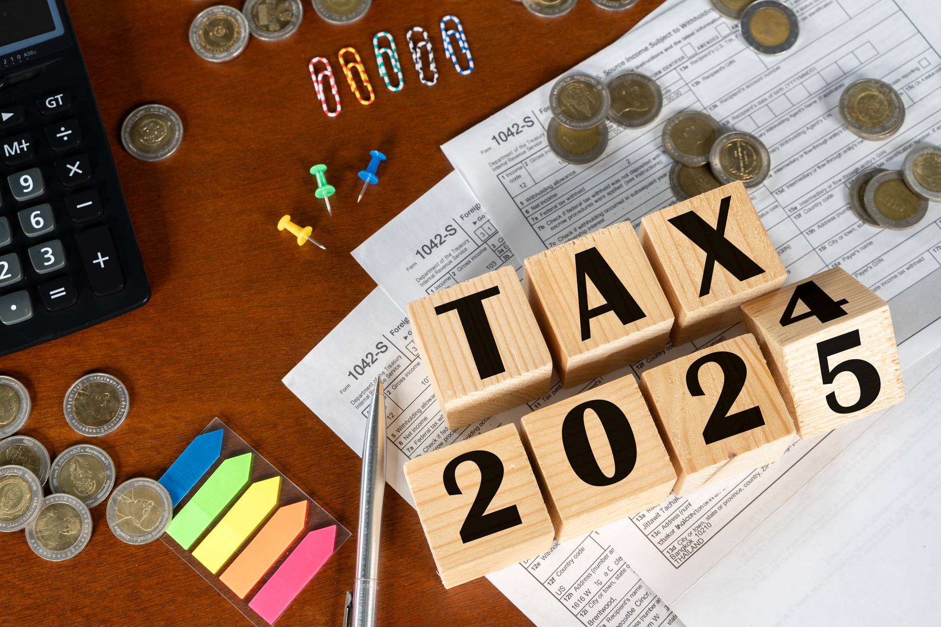 A wooden block with the word tax on it is on top of a pile of coins and a calculator.